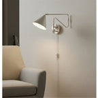 Fenwick Swing Arm Plug-In Wall Light in brushed steel, lit beside an armchair in a living room