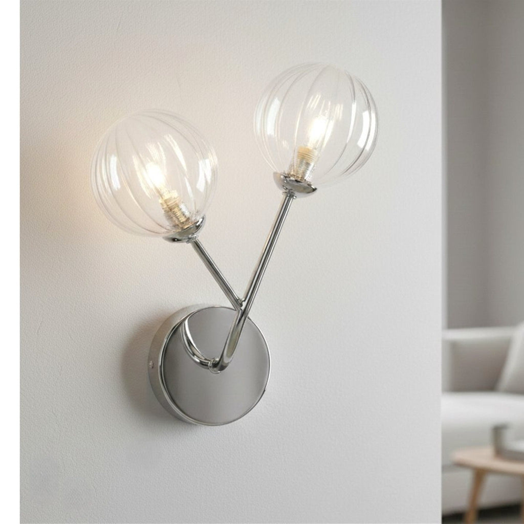 Lyra Twin Globe Wall Light in chrome lit on a pale wall beside a sofa