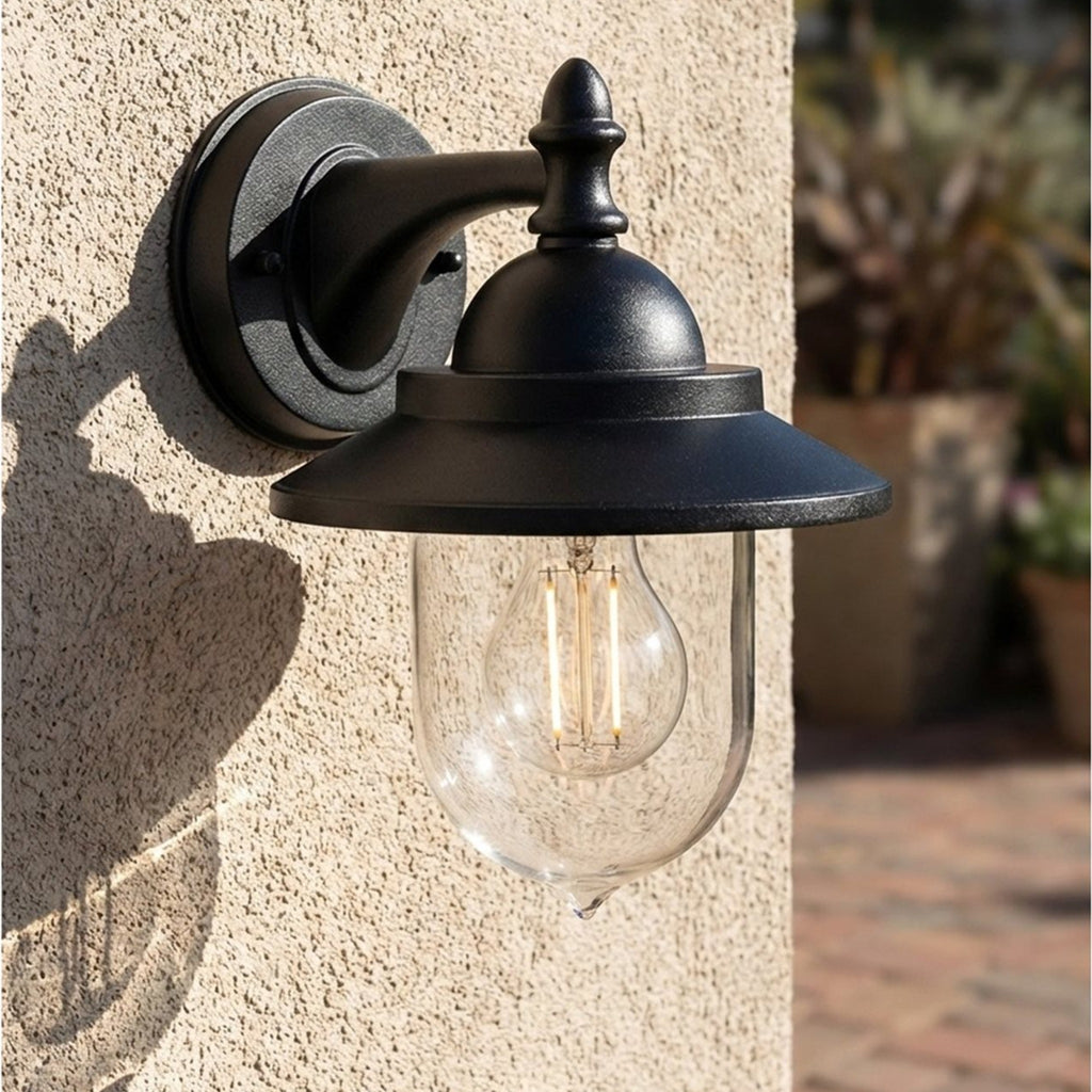 Padstow Clear Glass Outdoor Wall Lantern in black, lit and mounted on a rendered exterior wall beside a patio