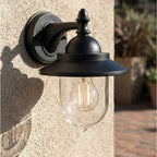 Padstow Clear Glass Outdoor Wall Lantern in black, lit and mounted on a rendered exterior wall beside a patio