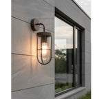 Tarbet Caged Glass Outdoor Wall Light in rustic brown, lit on a rendered exterior wall beside a window